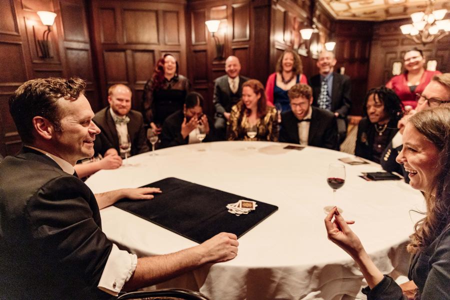 Guests sitting at a round table smiling and laughing while Dennis performs magic.