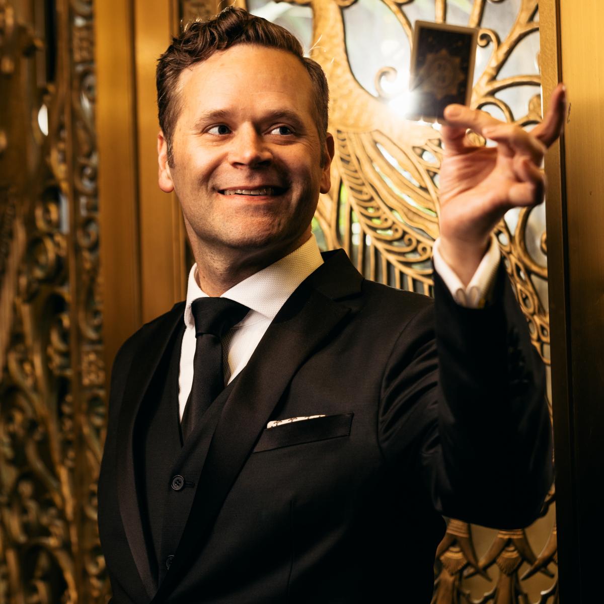 Dennis stands in front of gold doors with a playing card.